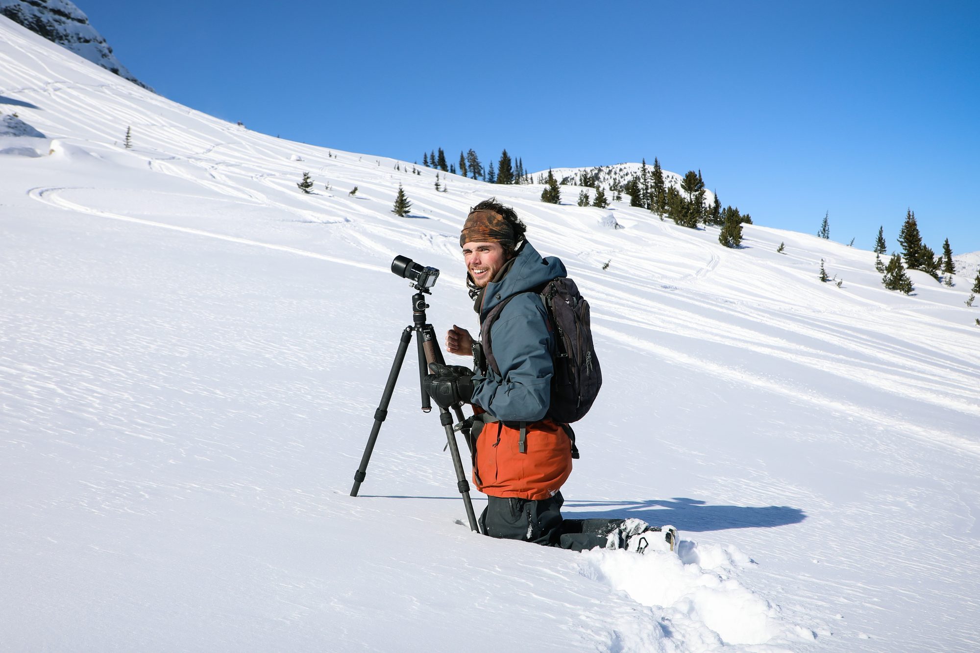 Jack kneeling in deep snow beside a camera on a tripod, grinning, on a sunny backcountry slope
