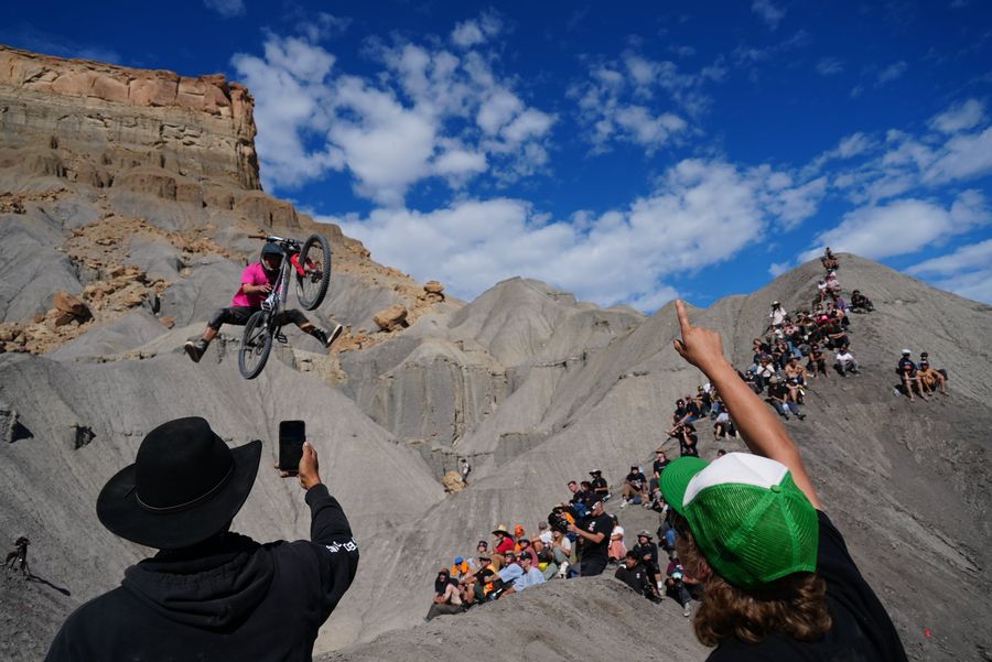 A mountain biker airborne over desert badlands while a crowd on the ridge cheers