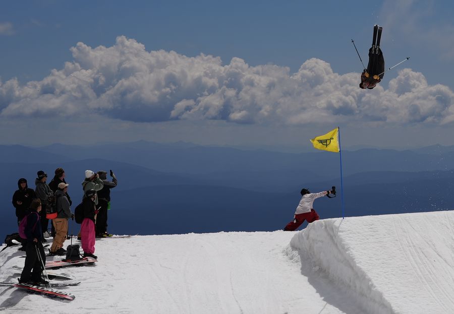 A skier flipping off a jump as a filmer and a group of riders watch from the snow