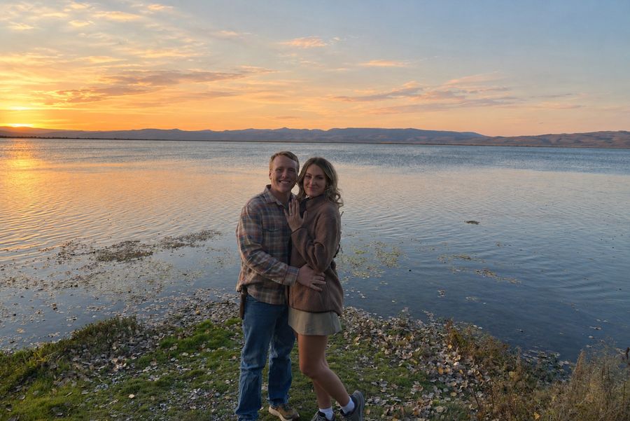 A newly engaged couple smiling by a lake at sunset, her ring hand on his chest