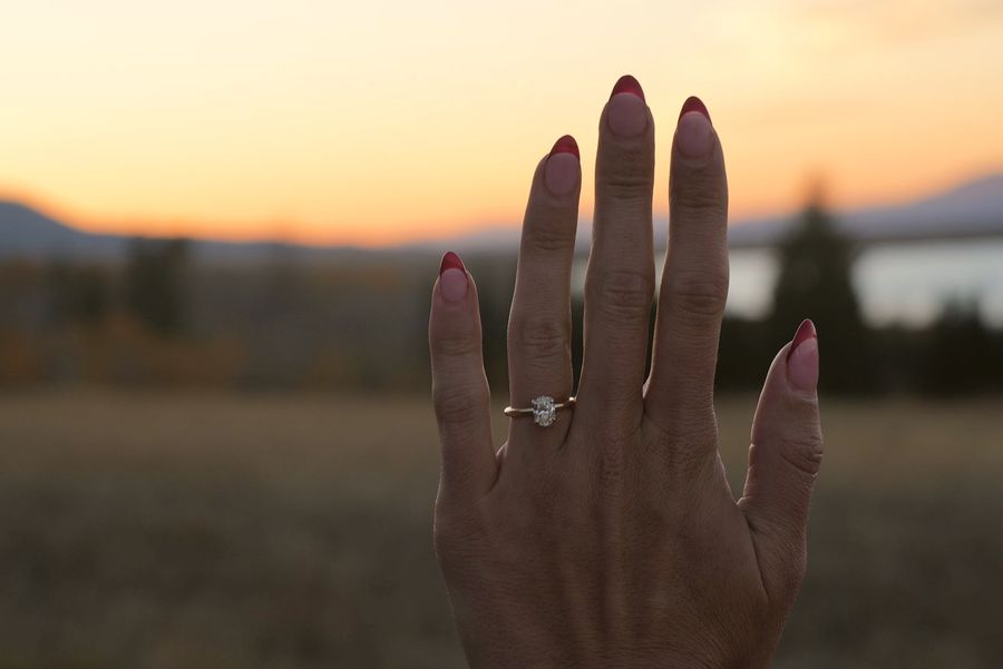 A hand with an oval solitaire engagement ring held up against a sunset over hills and water