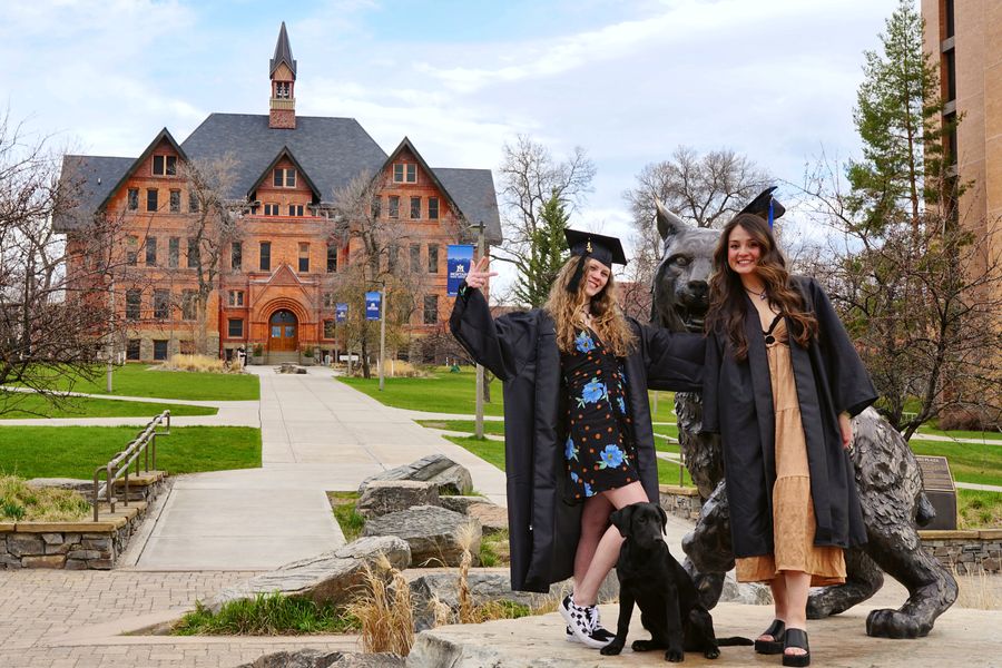 Two graduates in gowns with a black lab puppy beside the bronze bobcat statue at Montana State