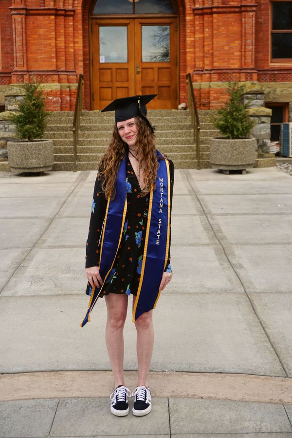 A graduate in a Montana State stole standing before the steps of a brick hall