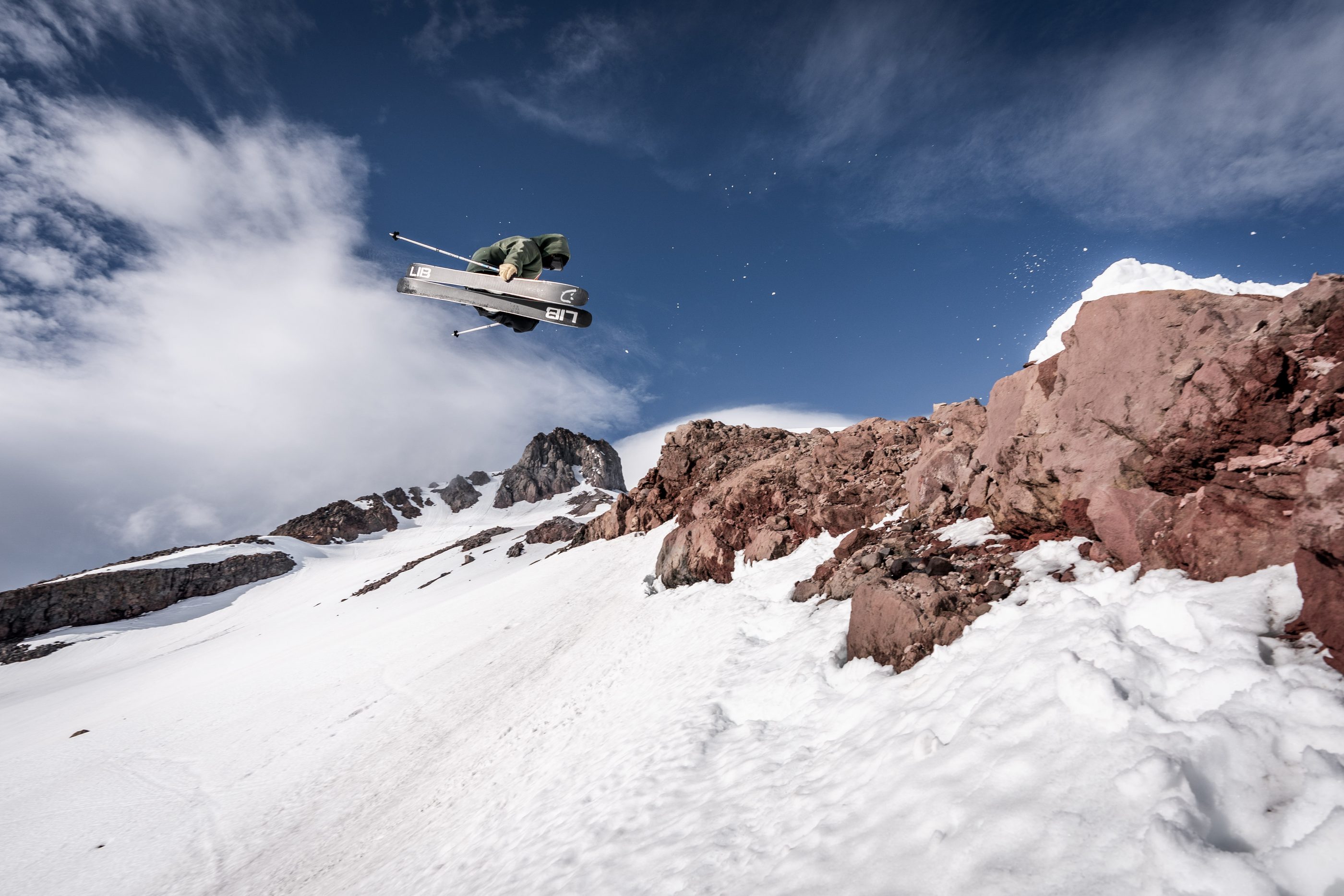 A skier airborne over a rocky, snow-covered ridgeline under a deep blue sky
