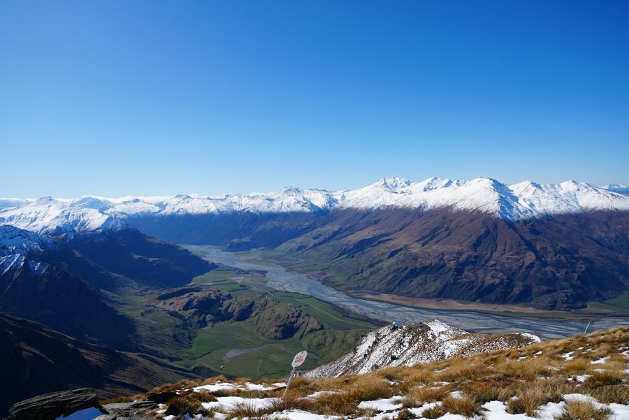 A braided river valley beneath a long snowy range, seen from a summit